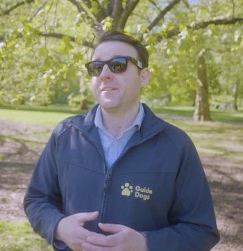 A photo of Tommy standing in a park wearing a Guide Dogs branded jacket and sunglasses.
