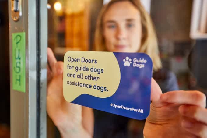 Woman places a High Street Heroes sticker by Guide Dogs into a window, letting guide dog and assistance dog owners know that the shop welcomes assistance dogs. 