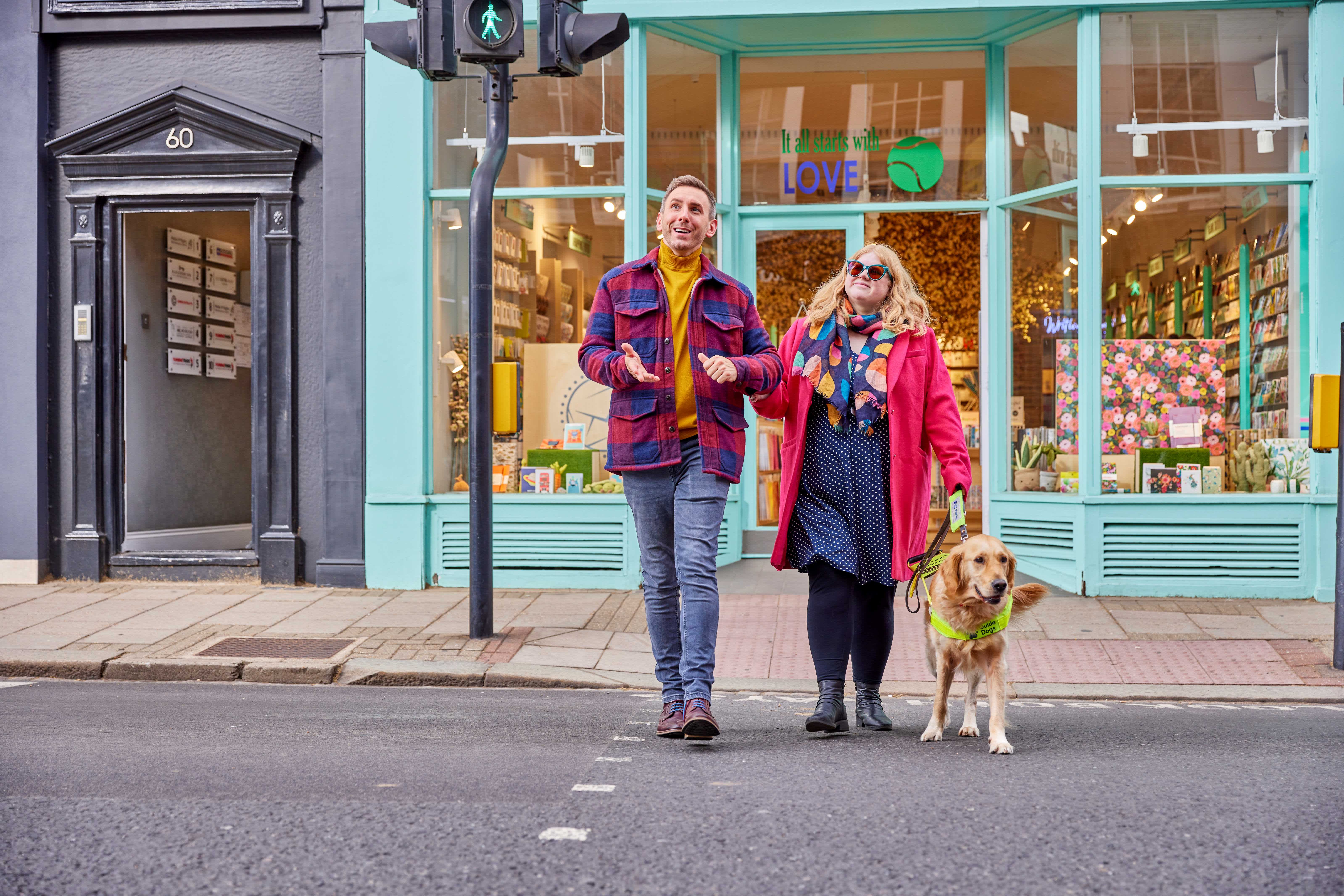 A person guides a guide dog owner across the road as they talk to each other and the guide dog walks beside them on the lead.