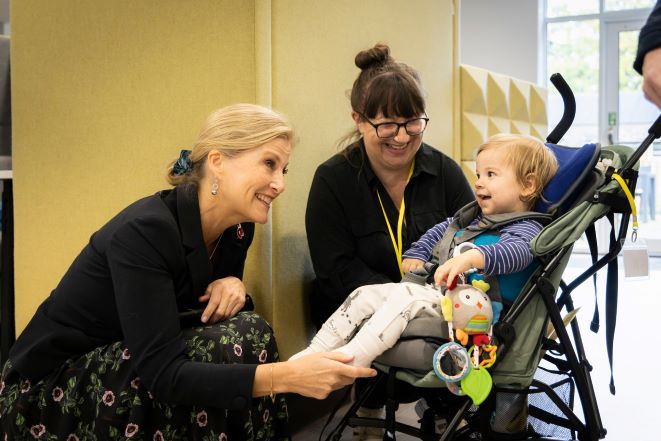 HRH The Duchess of Edinburgh crouches down to meet 1-year-old Theo in his pram and his mother Erika. Theo smiles at HRH while The Duchess and Erika smile back at Theo. 