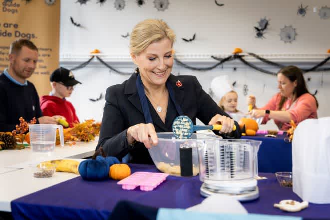 HRH The Duchess of Edinburgh making Halloween dog treats at a table. She is smiling as she holds a bowl and utensil to mix the treats. There's families and Halloween decorations in the background. 