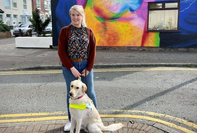 Lynette stands with her guide dog in front of a colourful building