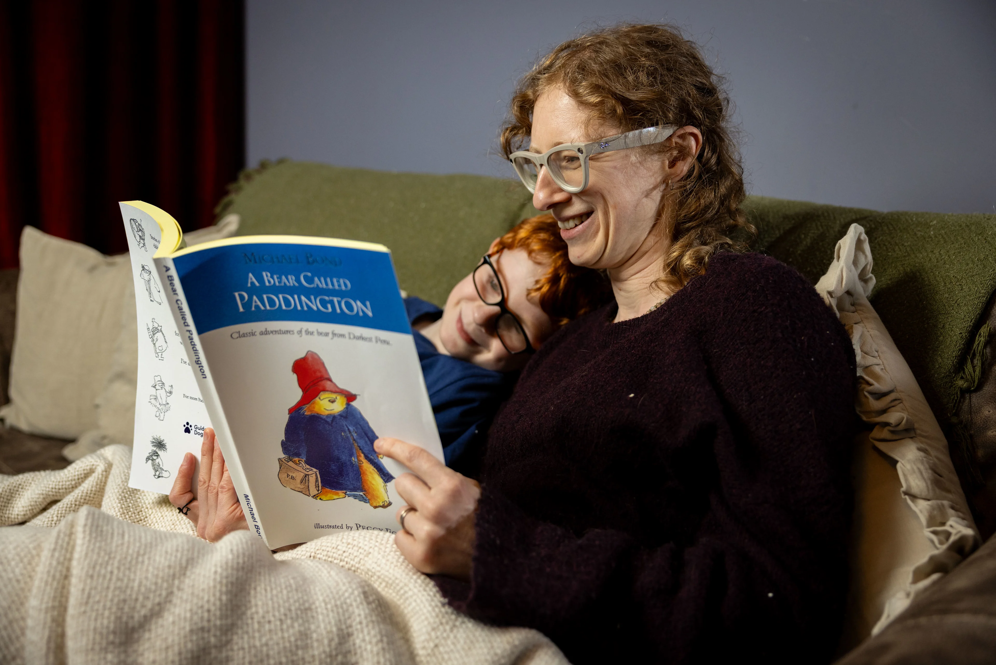 A mum reads 'A Bear Called Paddington' to her son on the sofa who is resting his head on her shoulder. 