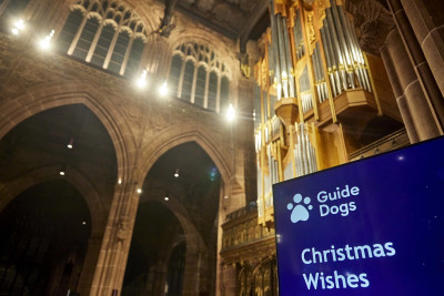 A blue Guide Dogs Christmas Wishes banner in the foreground in front of a grand view of the arches of Manchester Cathedral