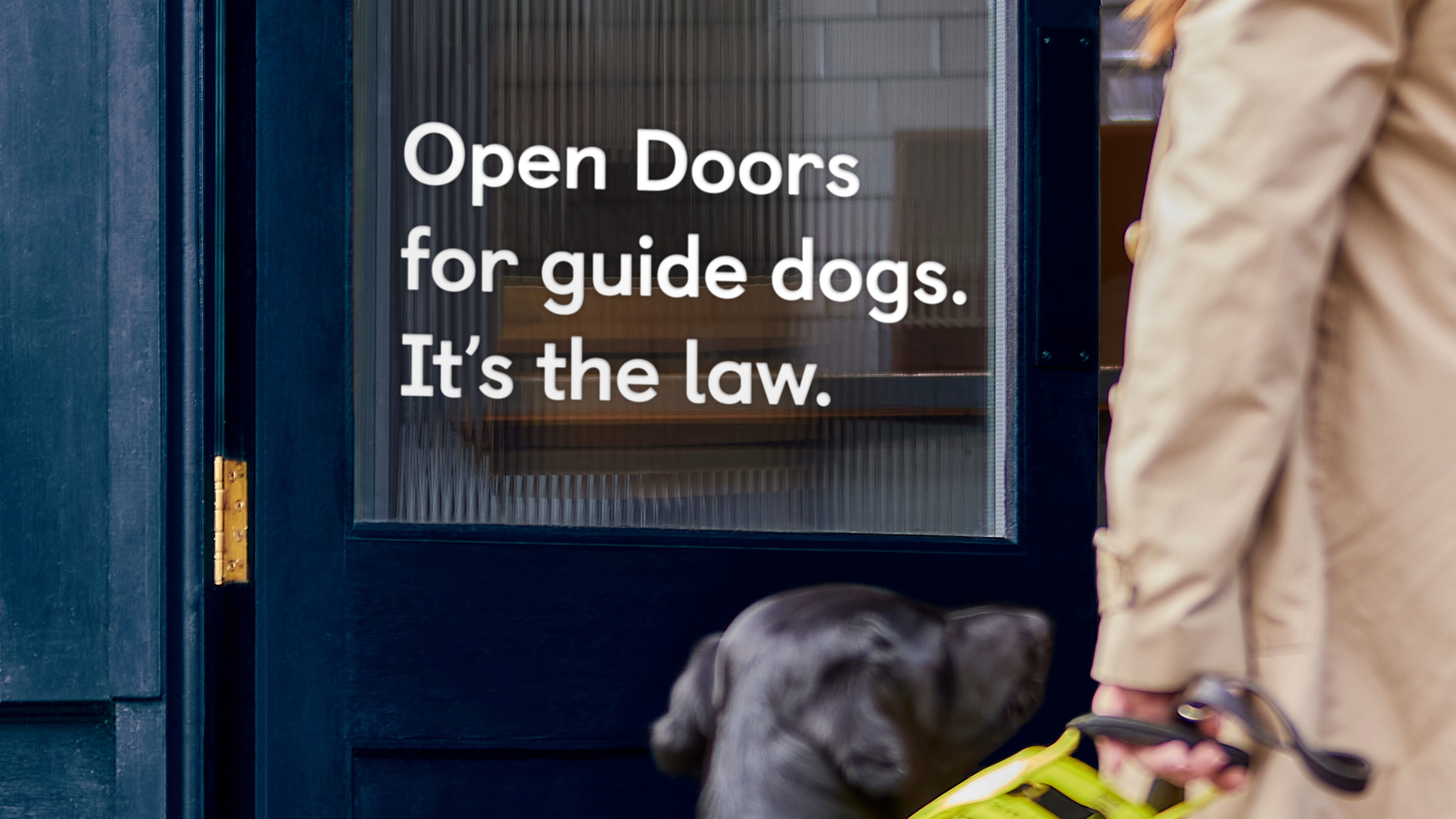 A guide dog owner and her black guide dog walk towards a door with a sign on the door saying "Open Doors for guide dogs. It's the law".