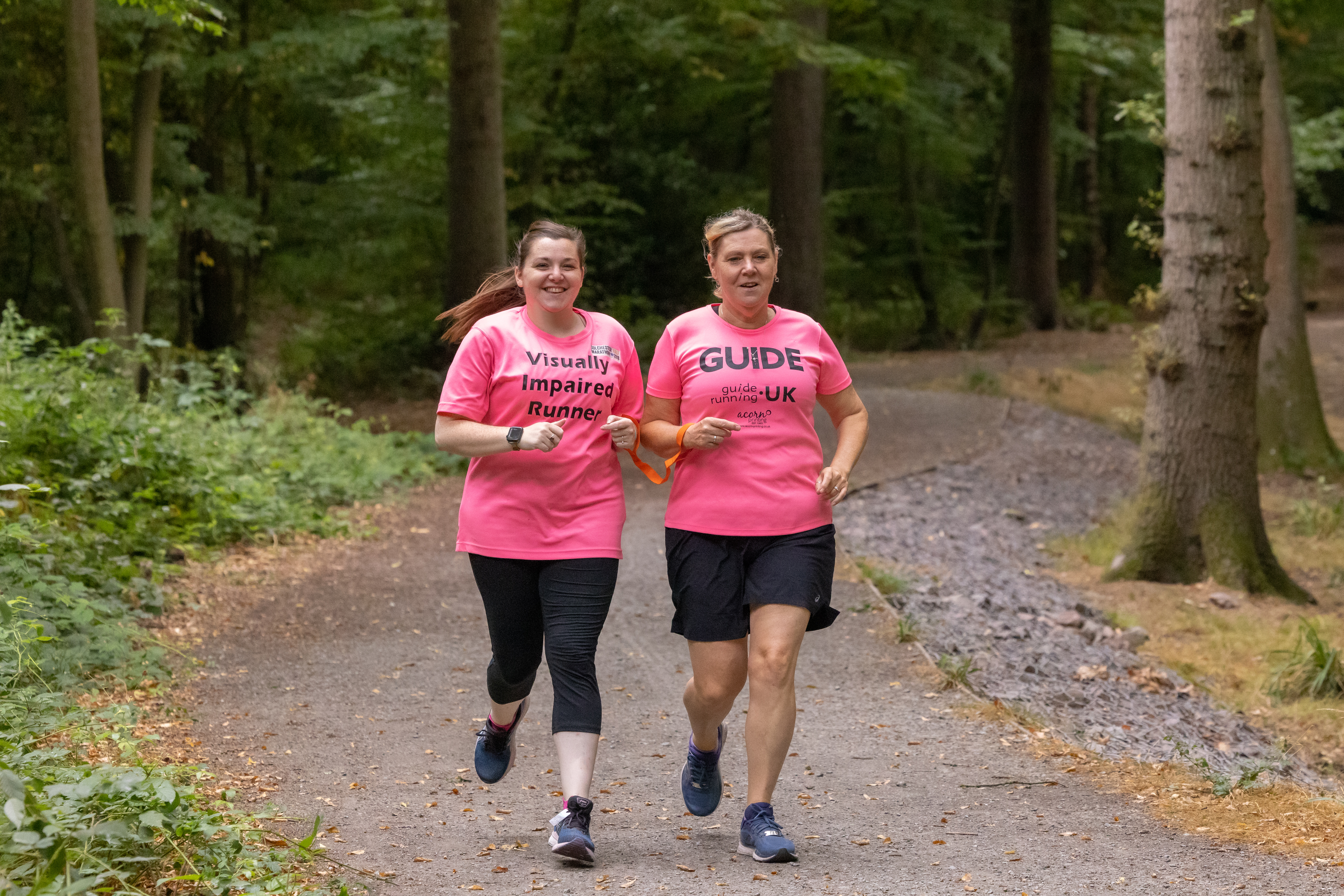 Samantha and Linda are running together through woods. They are both wearing pink t-shirts; Samantha's t-shirt says 'visually impaired runner' and Linda's t-shirt says 'Guide runner'