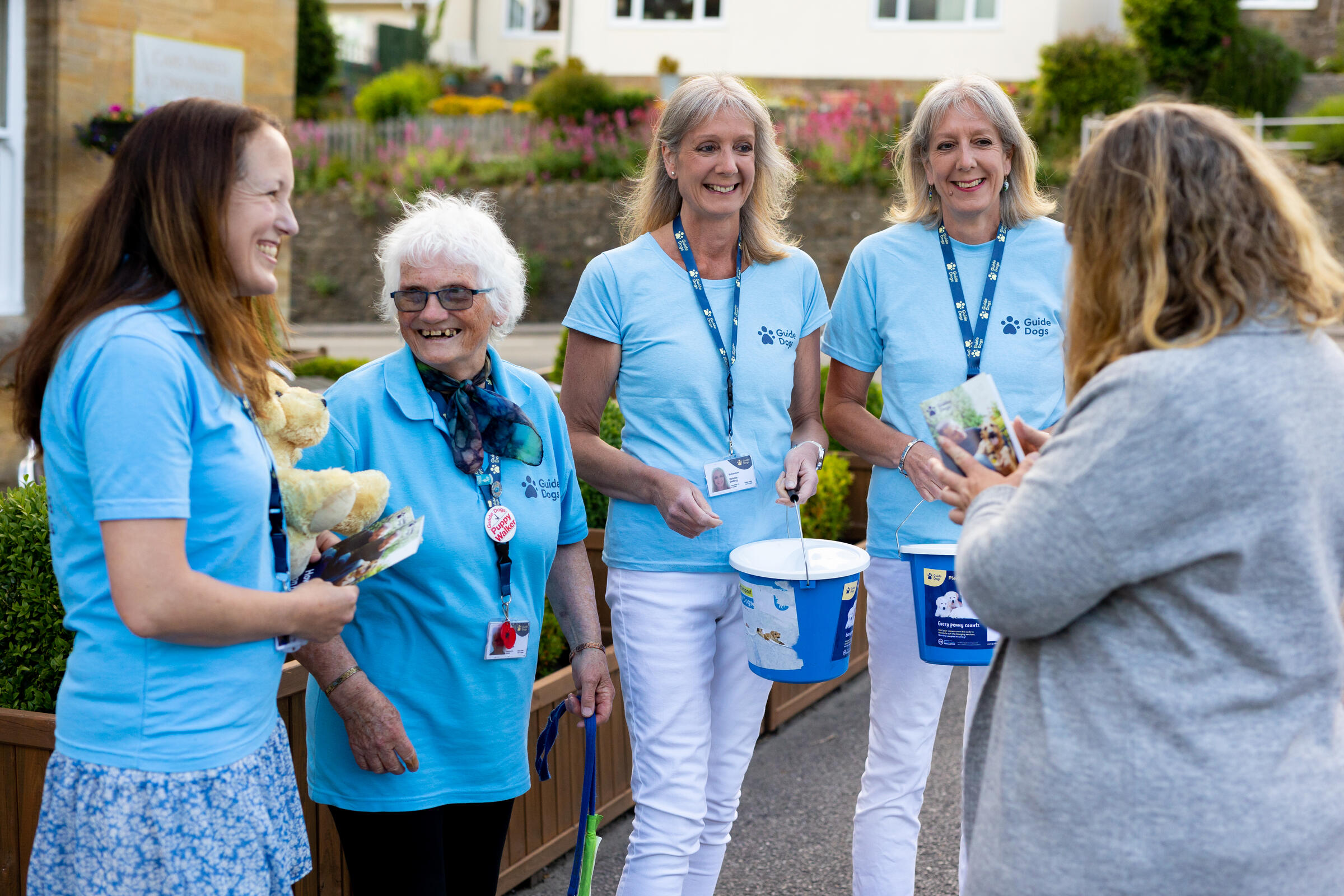 Four women stand in a row. They are smiling and wearing light blue Guide Dogs volunteer t-shirts. Two women hold collection buckets and one holds a guide dog puppy toy. The volunteers are talking to a member of the public.