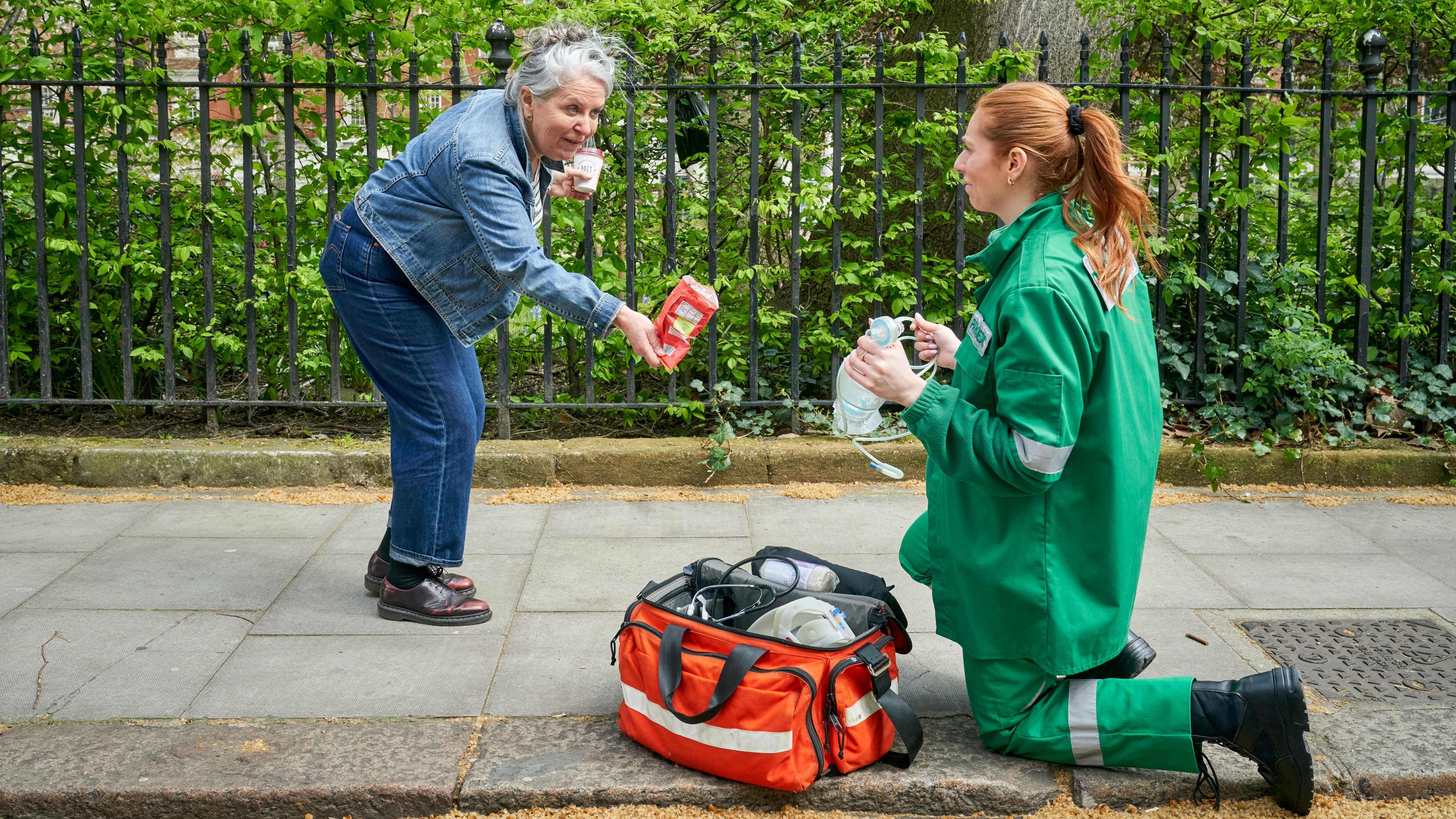 A paramedic in a green uniform kneels in the street with an emergency medical bag, while a strange woman bends to her to offer her a crisp.