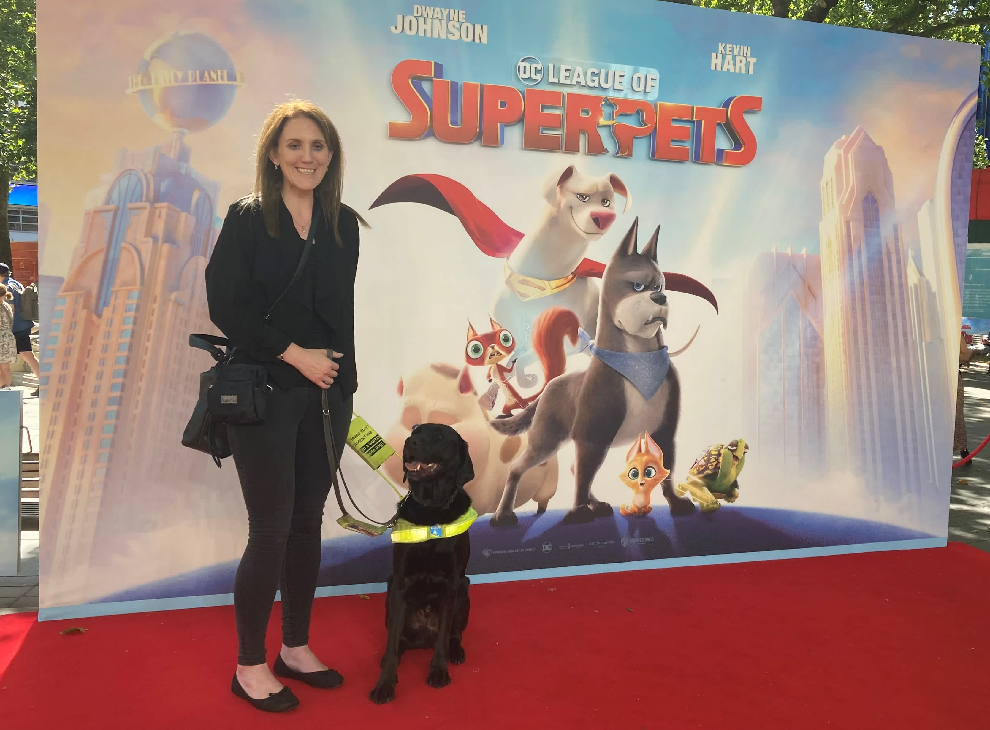 Siobhan and guide dog Marty stand on a red carpet stage in front of a poster for the new DC League of Super-Pets film. Siobhan is smiling with Marty on the lead to her left. They are standing on the left side of the poster so you can see the film's characters posing as a group in the centre and looking at the camera under the title 'DC League of Super-Pets'. 