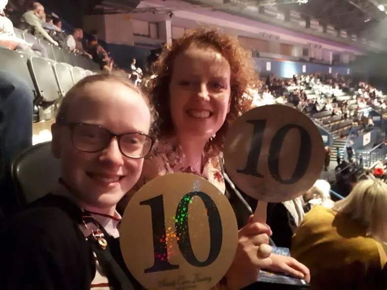 Neve sits in an audience with a lady as they both turn to smile towards the camera and hold up number 10 score cards. 