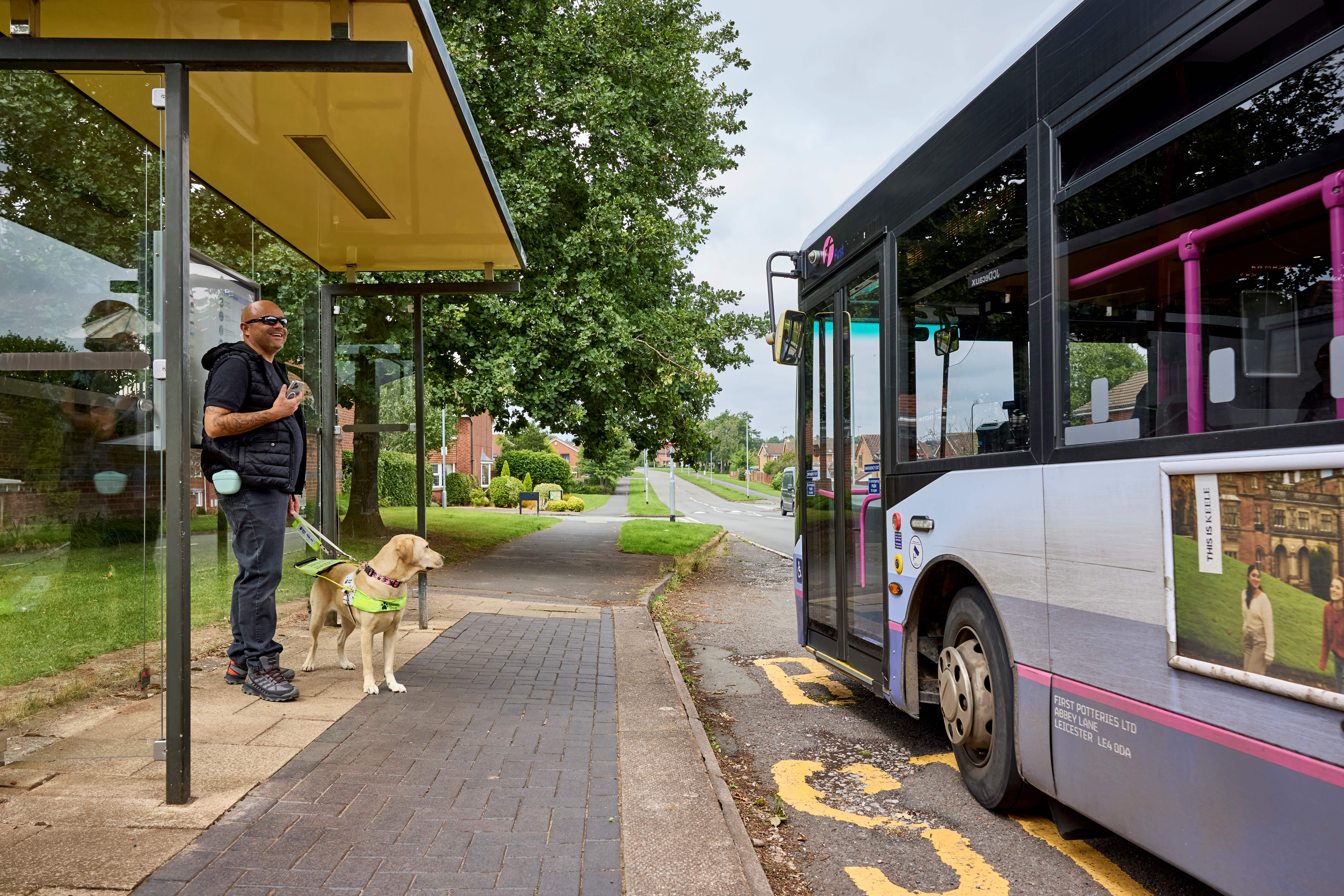 Guide dog owner stands at a bus stop with a guide dog in harness as a bus pulls into the bus stop. 