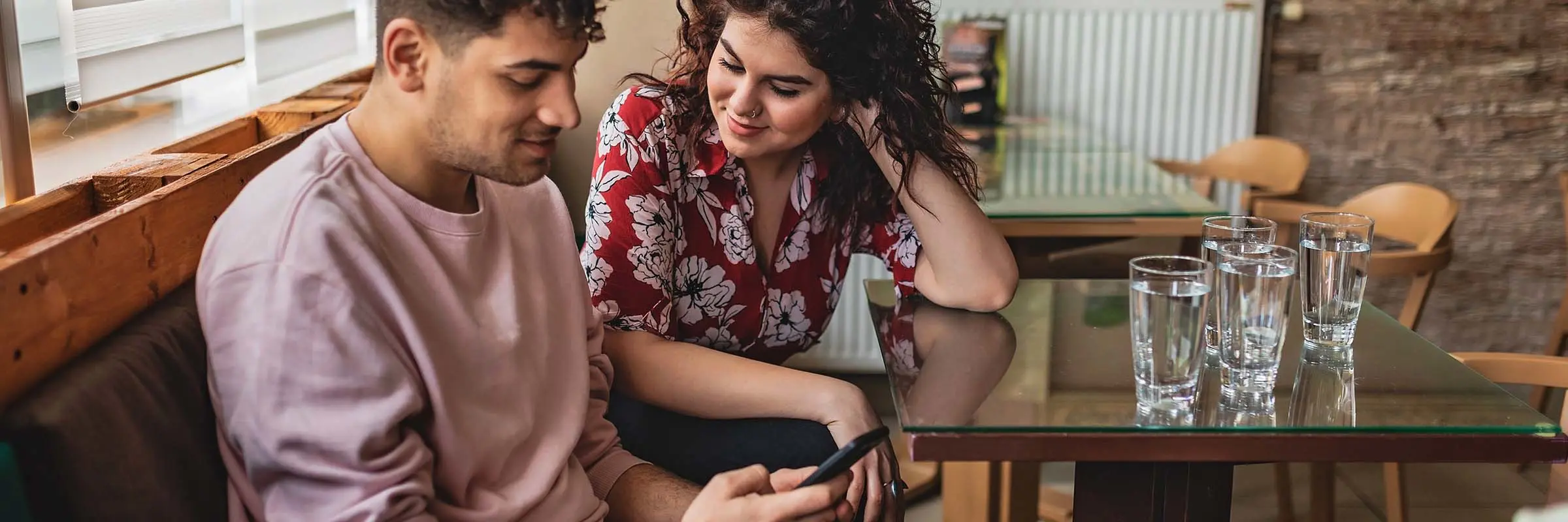 Man and woman sit together on a bench in a caf&eacute;. The man is looking down at his phone.