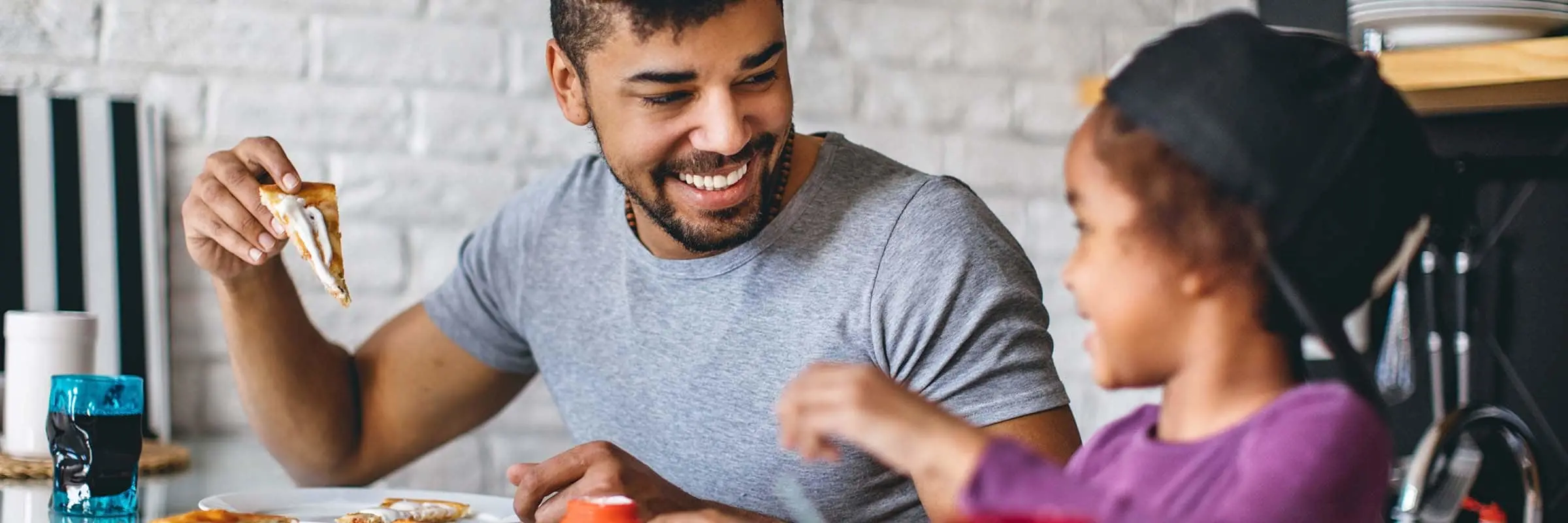 A father and daughter are smiling while eating pizza together in their kitchen.