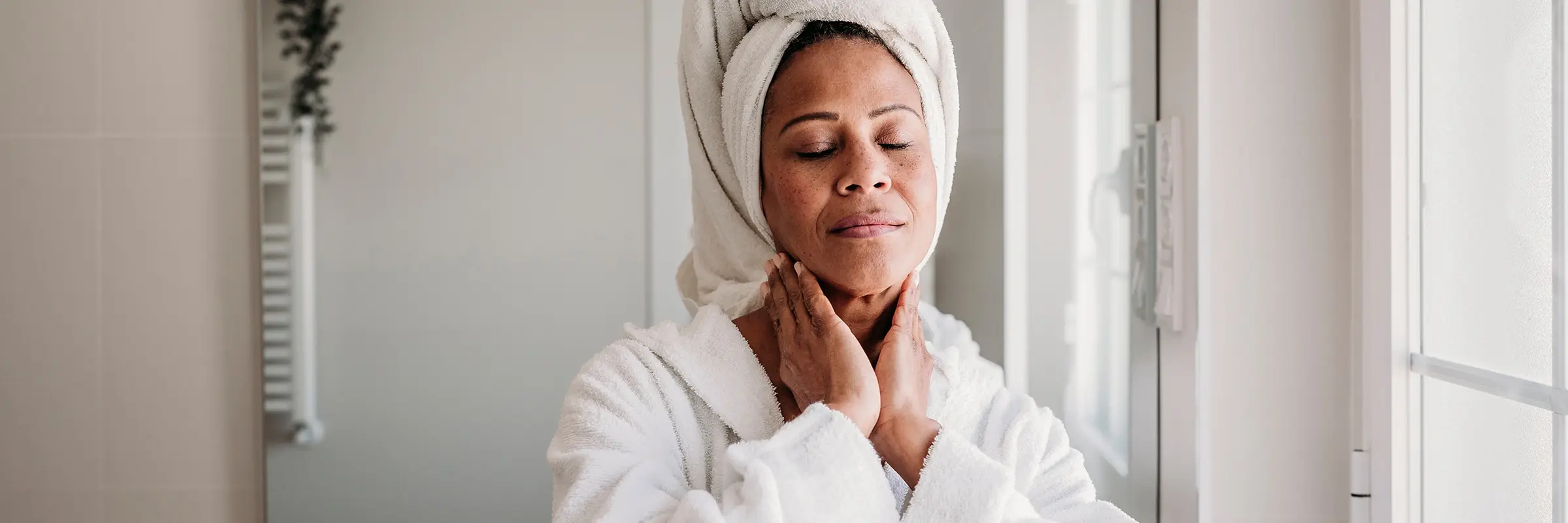 A woman rubbing her neck while wearing a robe with her hair wrapped in a towel.