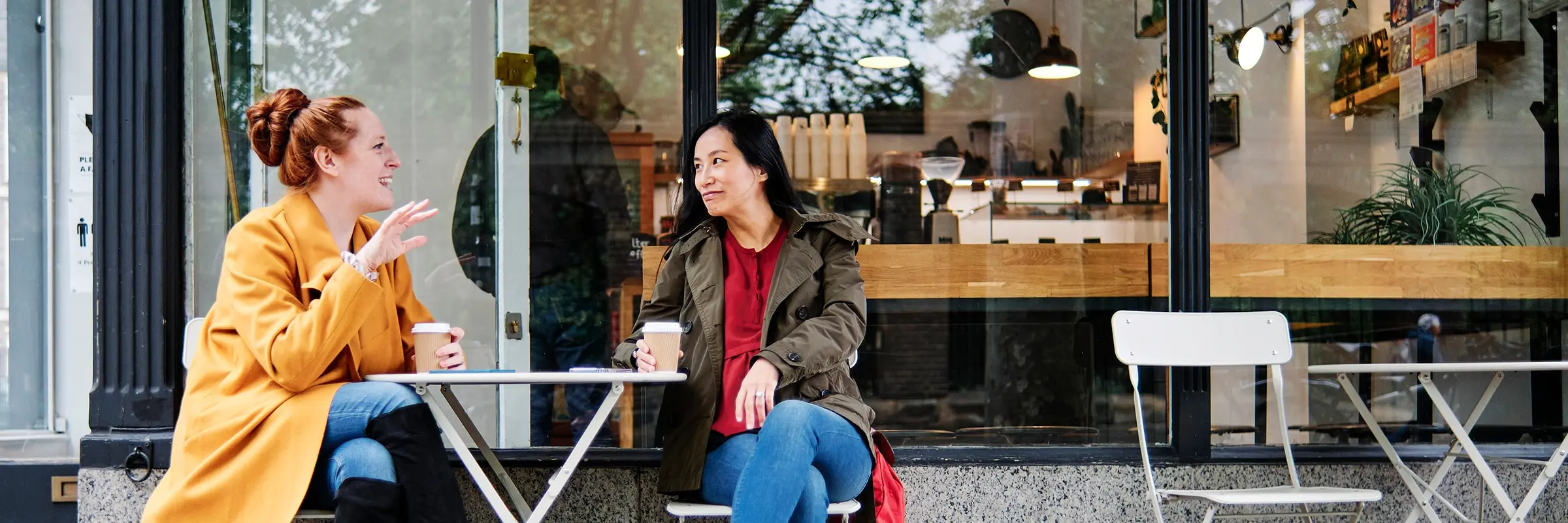 Two women drink coffee and chat outside of a cafe