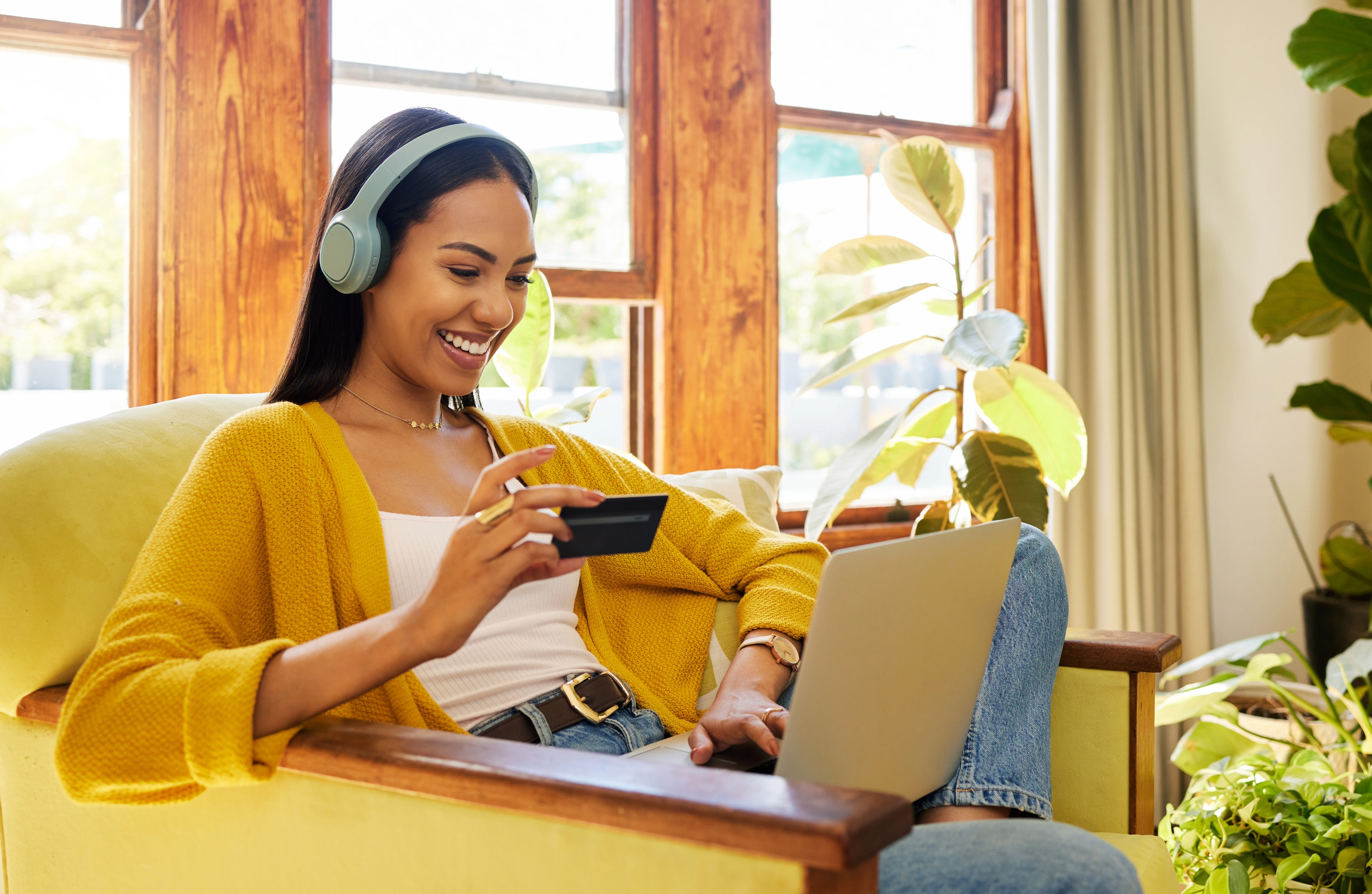 woman with headphone in a yellow sweater shopping on her laptop holding a credit card