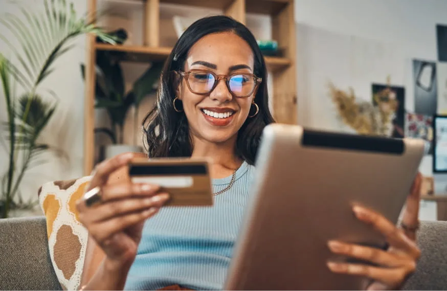 woman wearing glasses smiling down at a tablet while holding a credit card