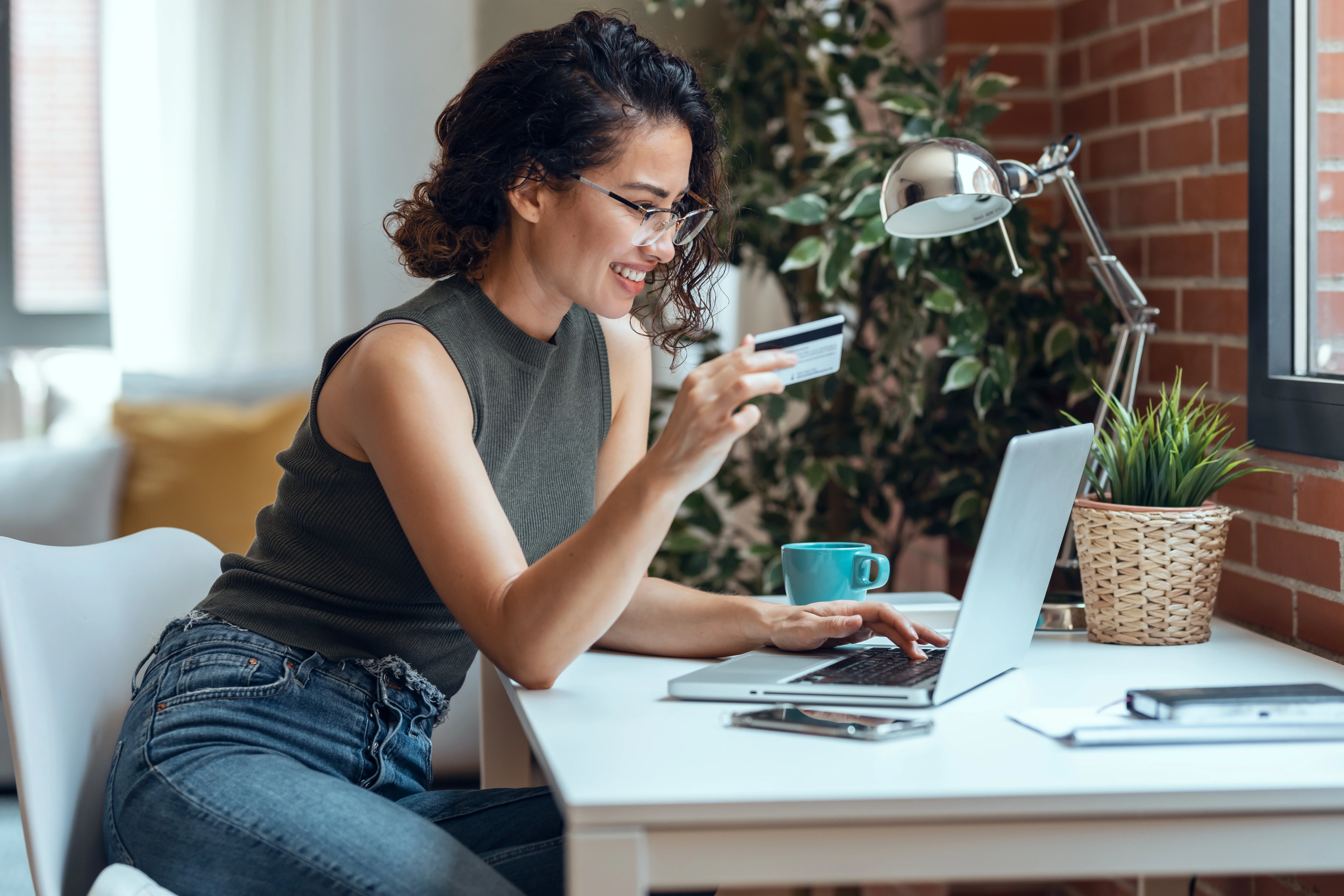 woman in glasses smiling while on her laptop and looking at her credit card