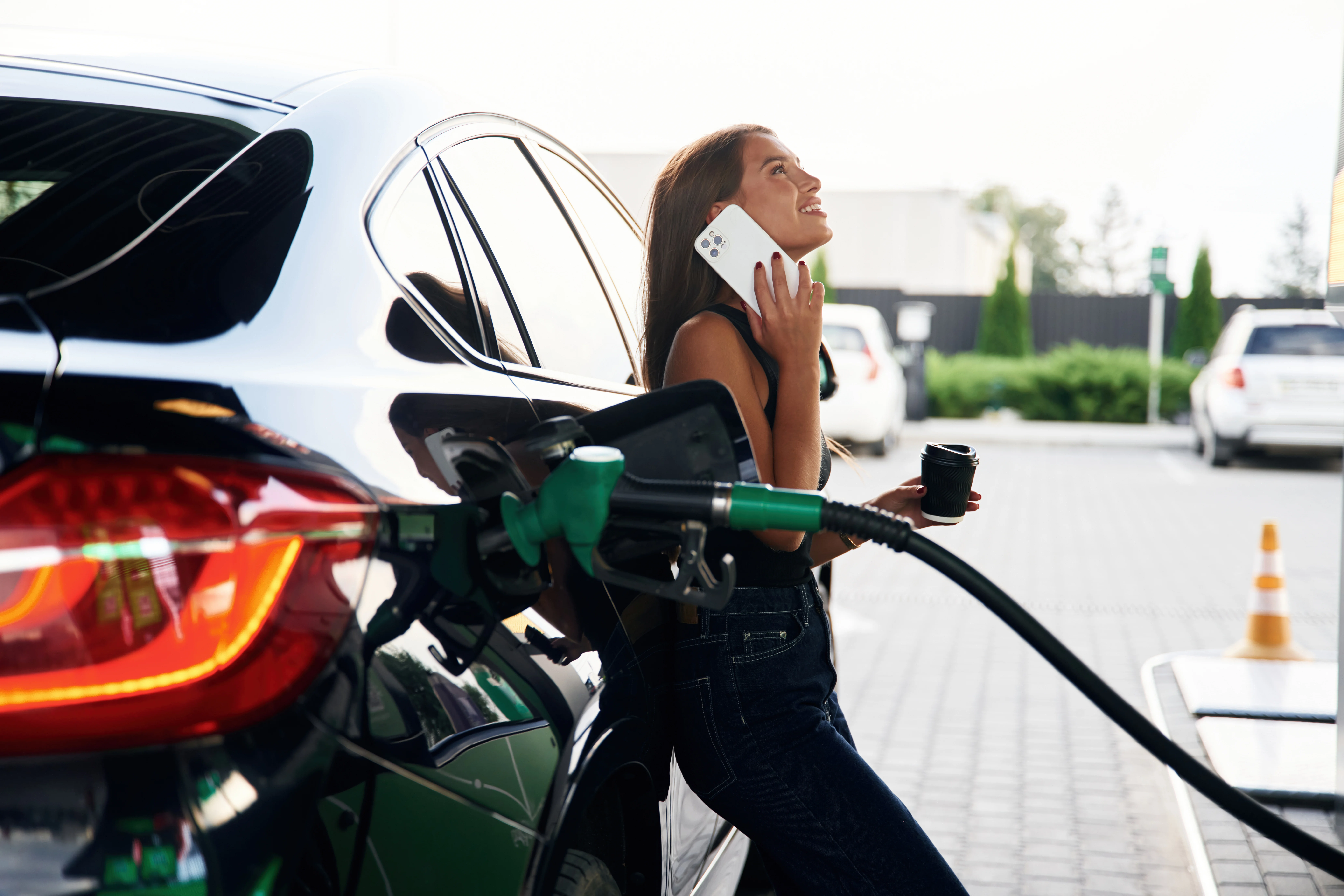 woman on phone with coffee leaning against her car while pumping gas