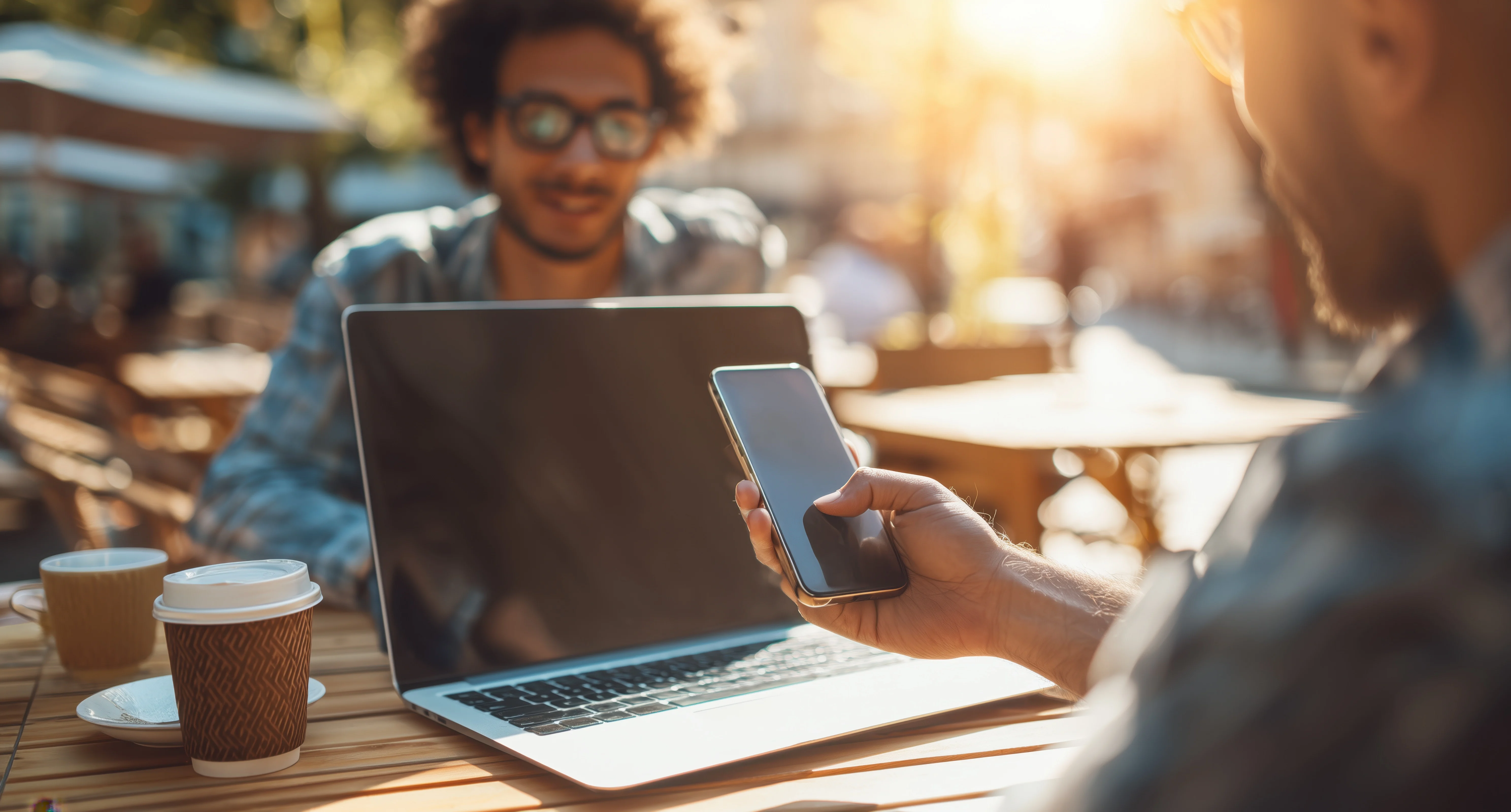 person holding their phone up sitting at table in front of laptop