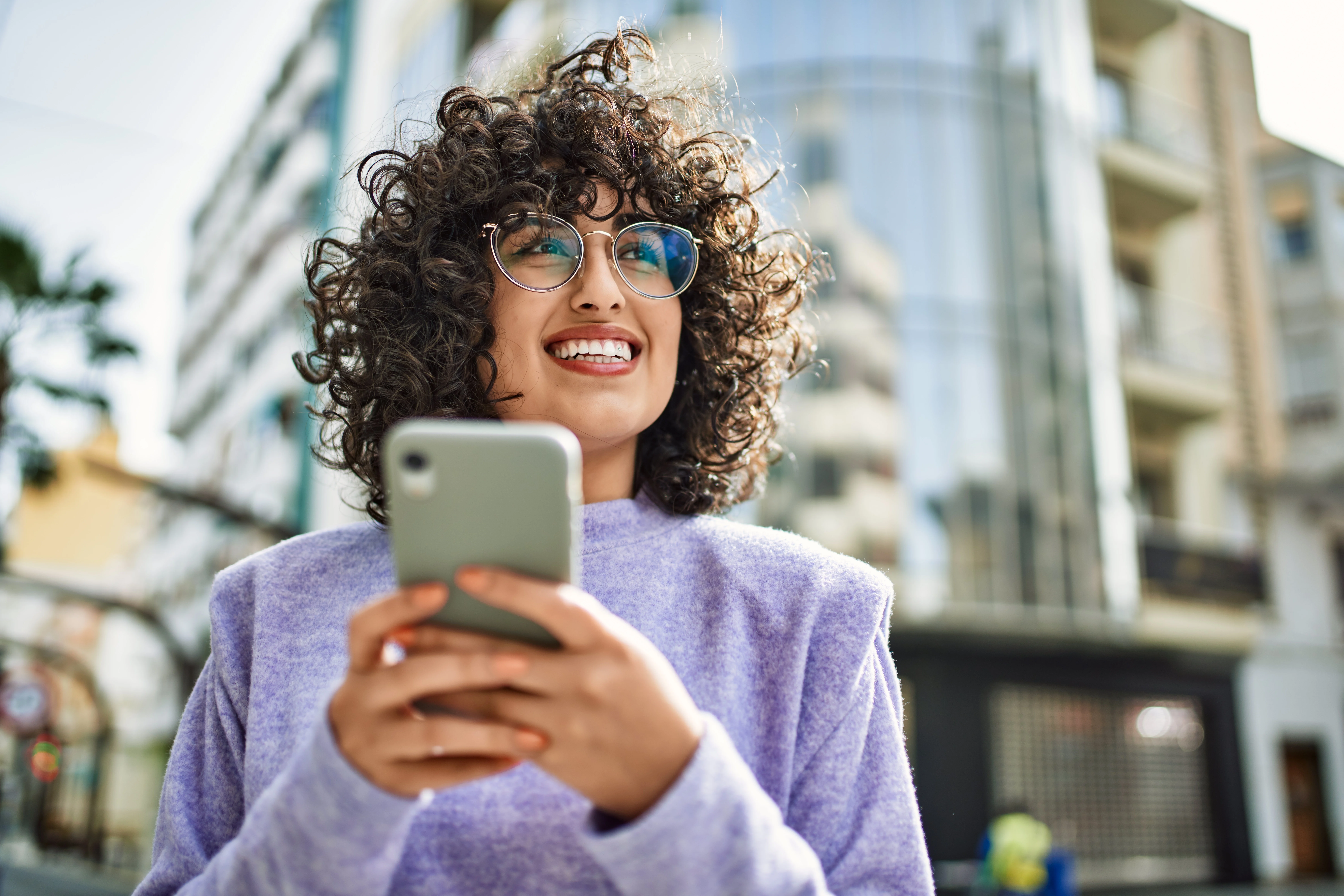 woman with glasses smiling, looking up while using her phone
