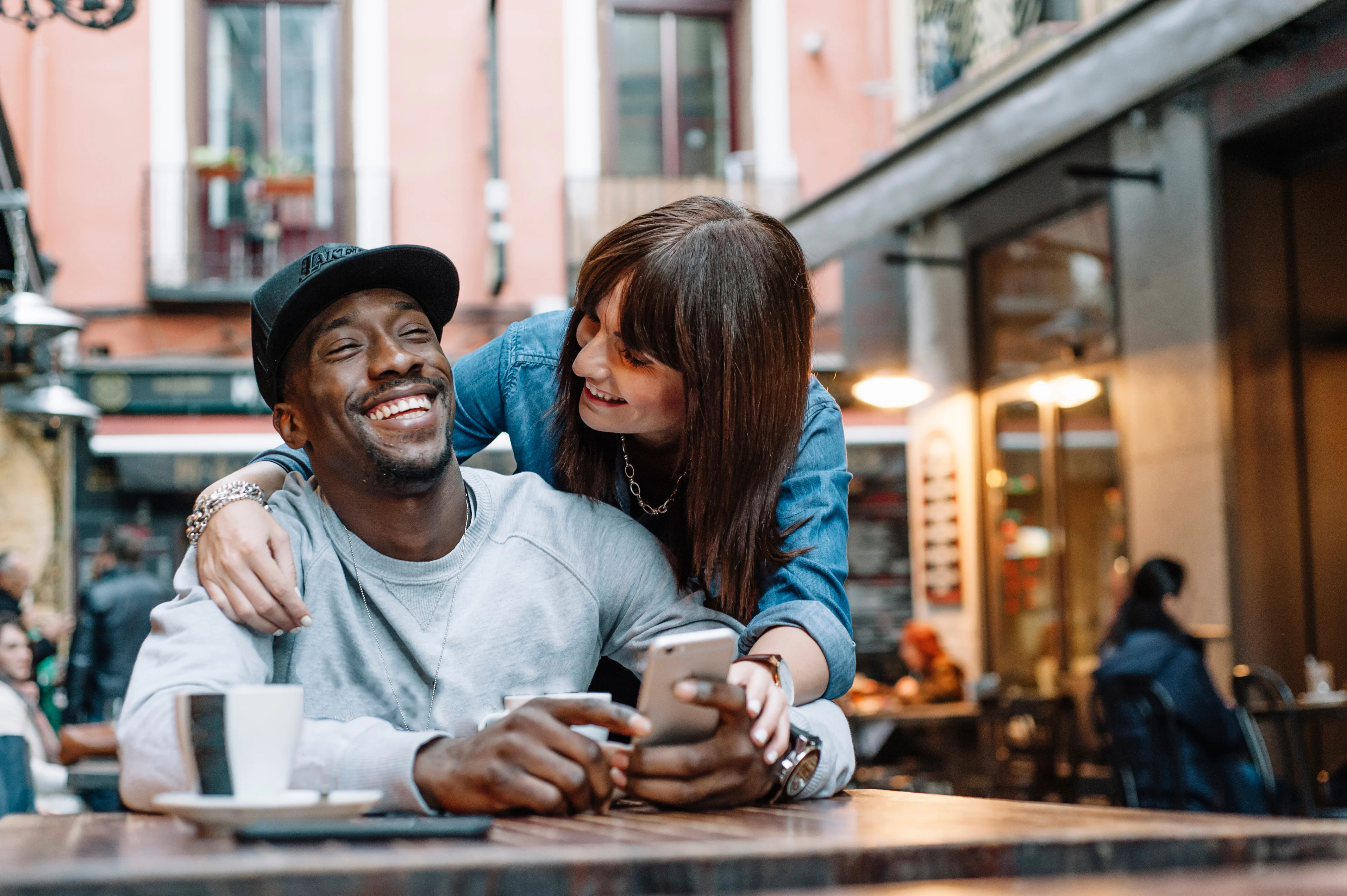 woman leaning over a man who is looking at his phone and hugging him and smiling