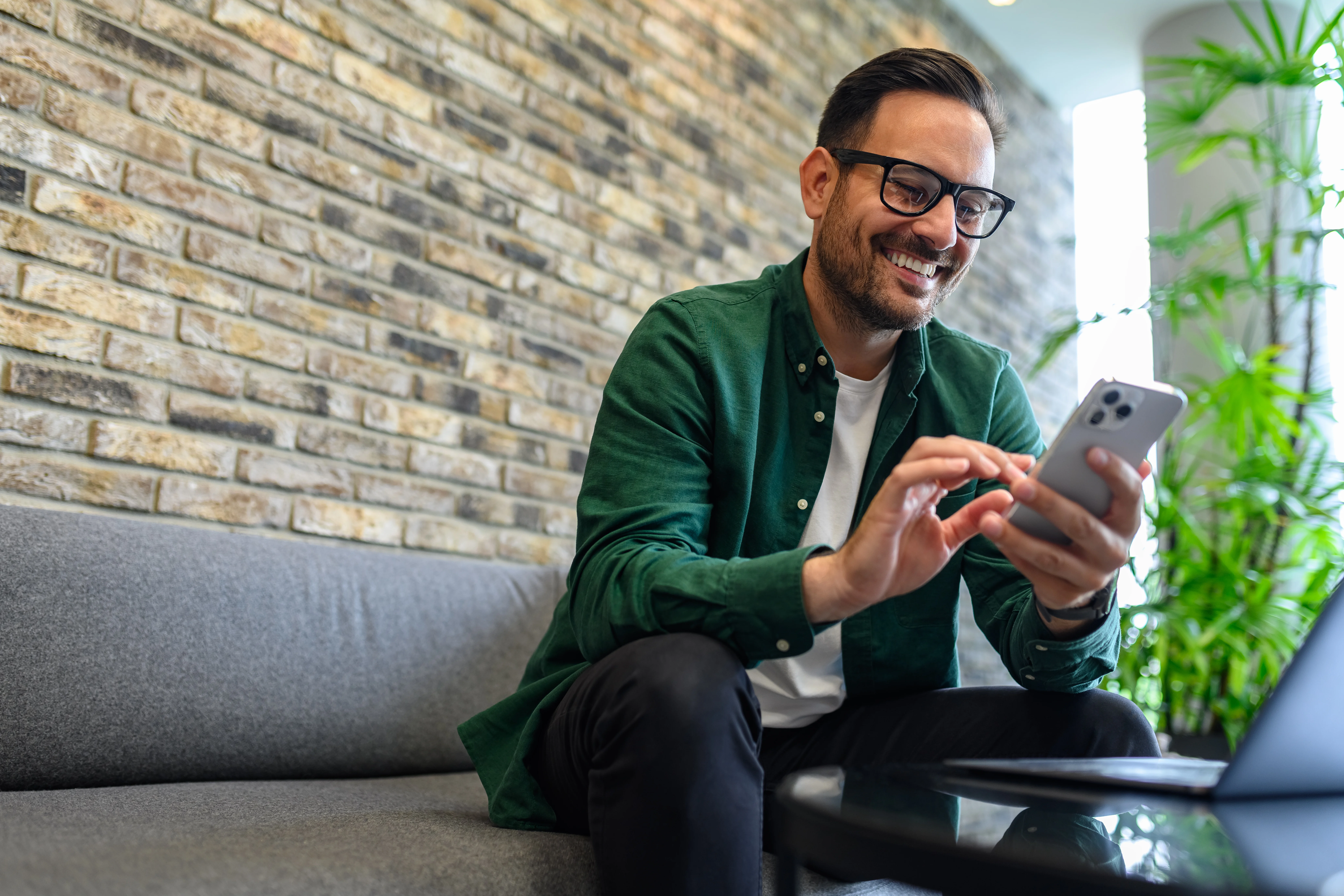 man smiling sitting on a couch with his phone and a plant in the background
