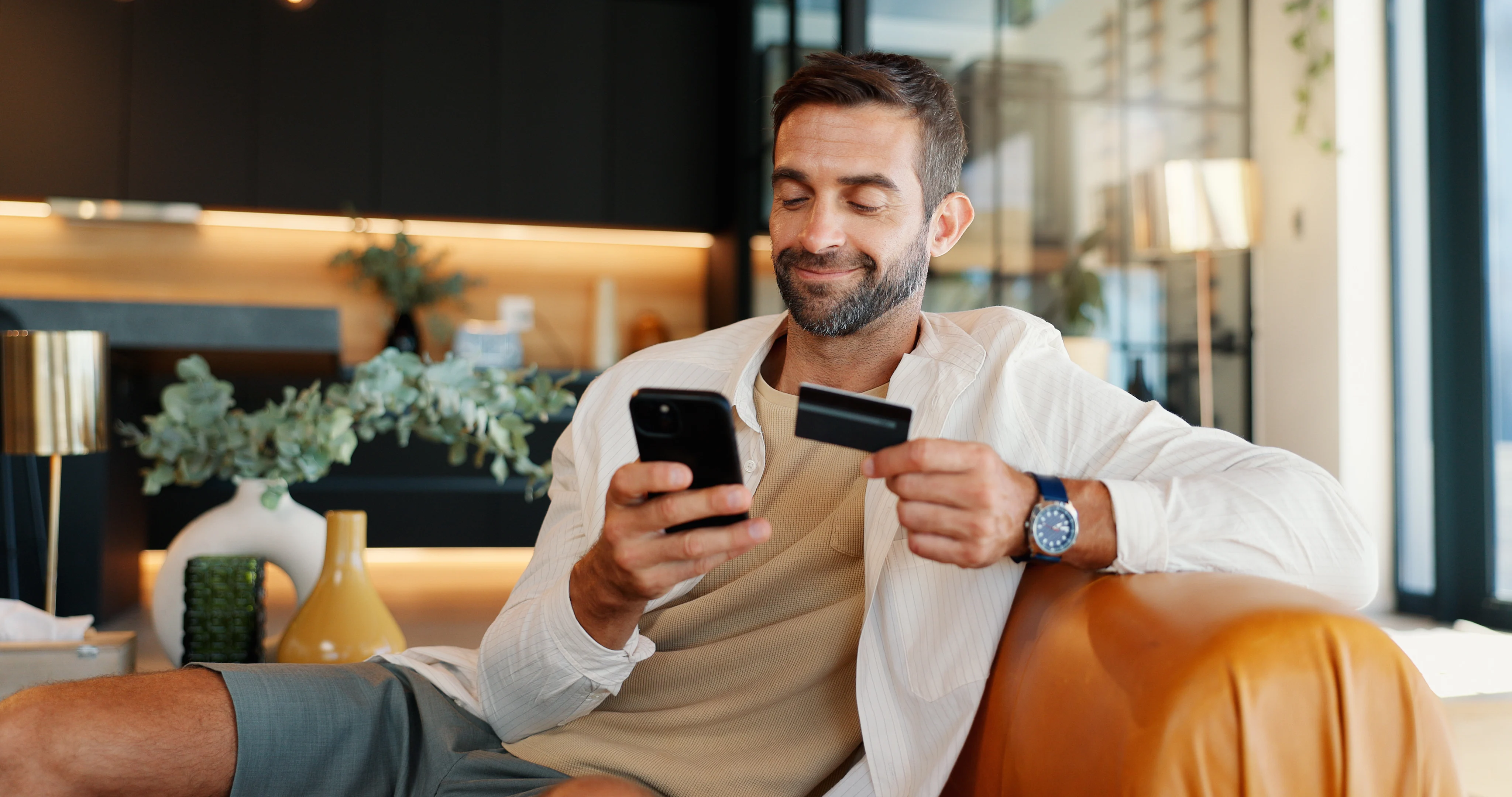 man sitting on couch smiling at phone while holding credit card