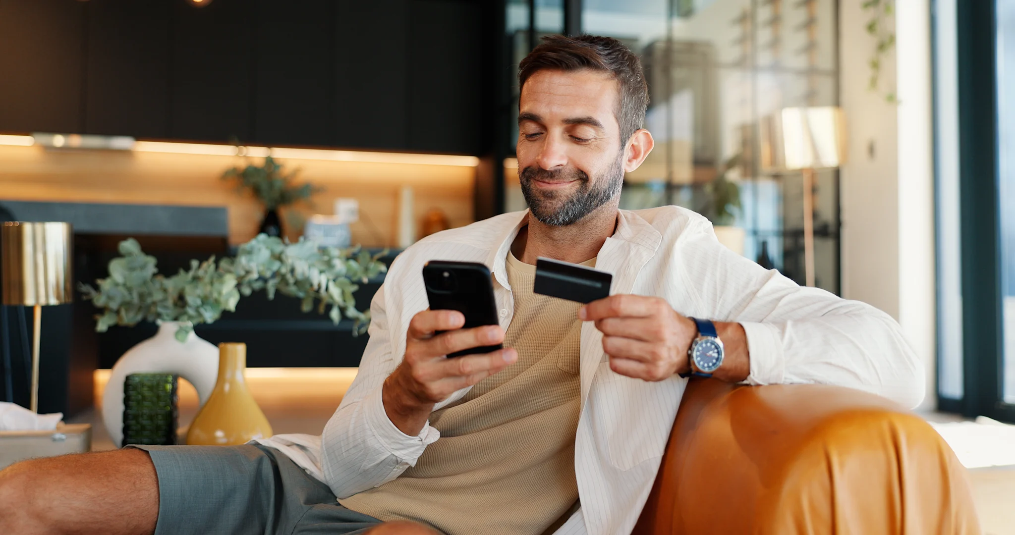 man sitting on couch smiling at phone while holding credit card