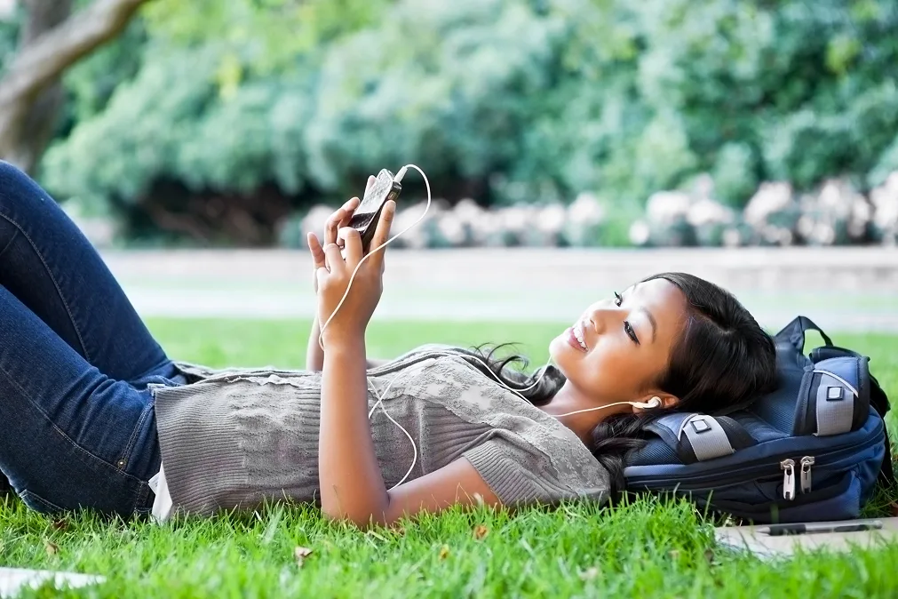 Mädchen, das mit einem Smartphone in der Hand und Kopfhörern in den Ohren auf dem Rasen liegt