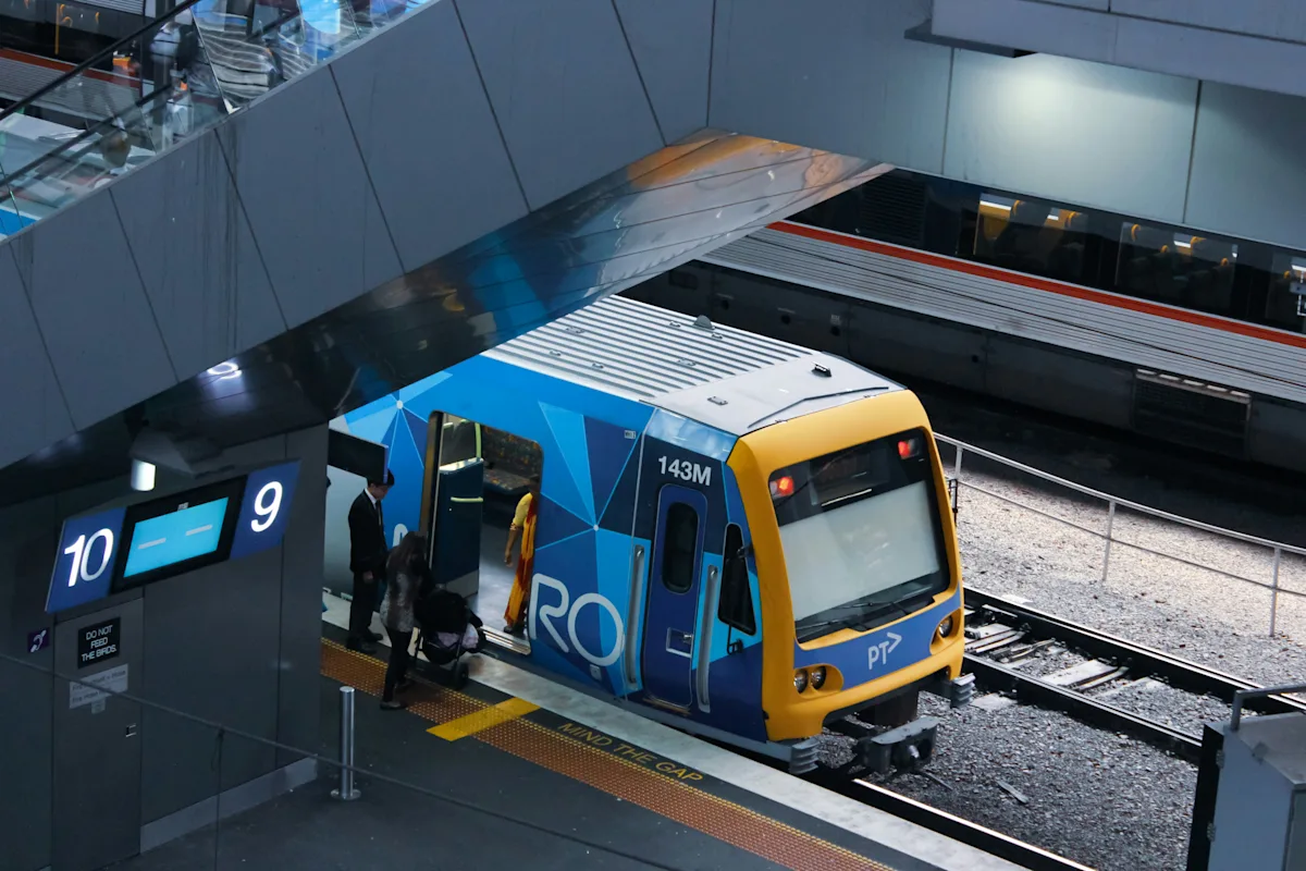 A blue and yellow Metro train stopped at a station platform in Melbourne, seen from above, with people boarding. Photo by Nao Takabayashi on Unsplash.