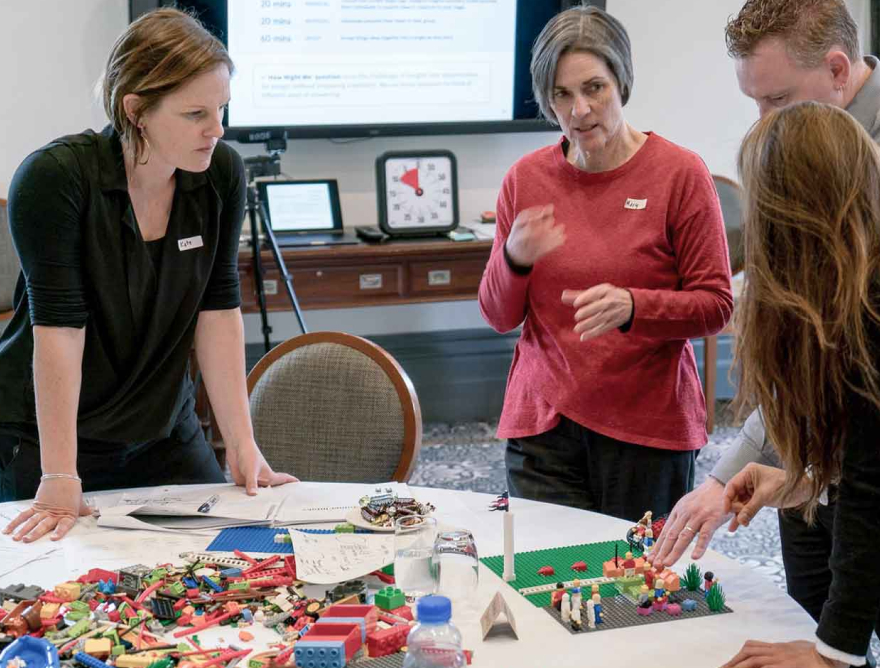 A group of four people are standing around a table covered in LEGO. One person is pointing to a scene made from LEGO while another is talking. They are all listening to the person talking. 