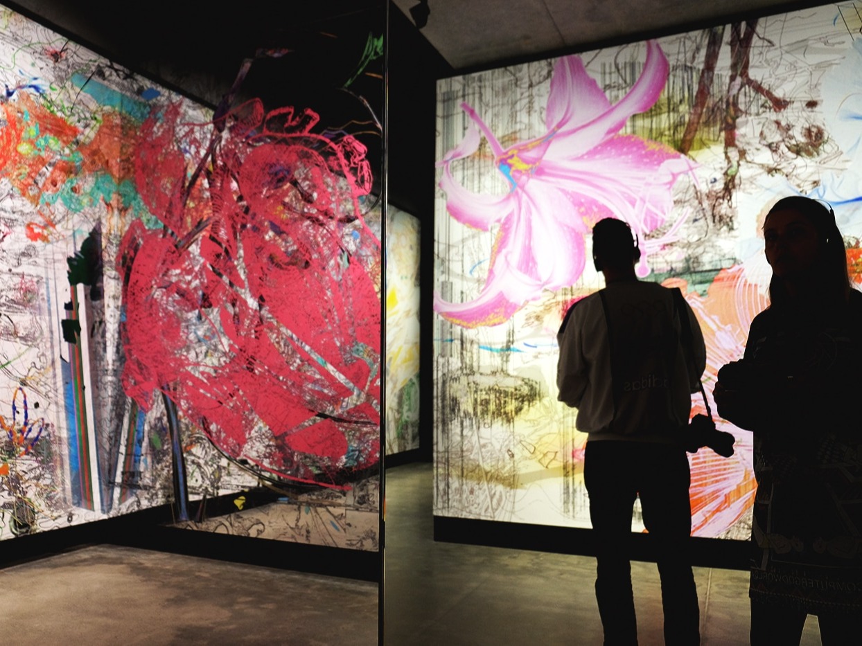 There are silhouettes of two people. They are walking through a gallery, that has two colourful illuminated panels with flowers and hand drawn sketches all over them.