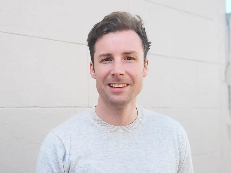 A photo of Jack Fahy smiling. He's outside in front of brick wall and it's a bright day.