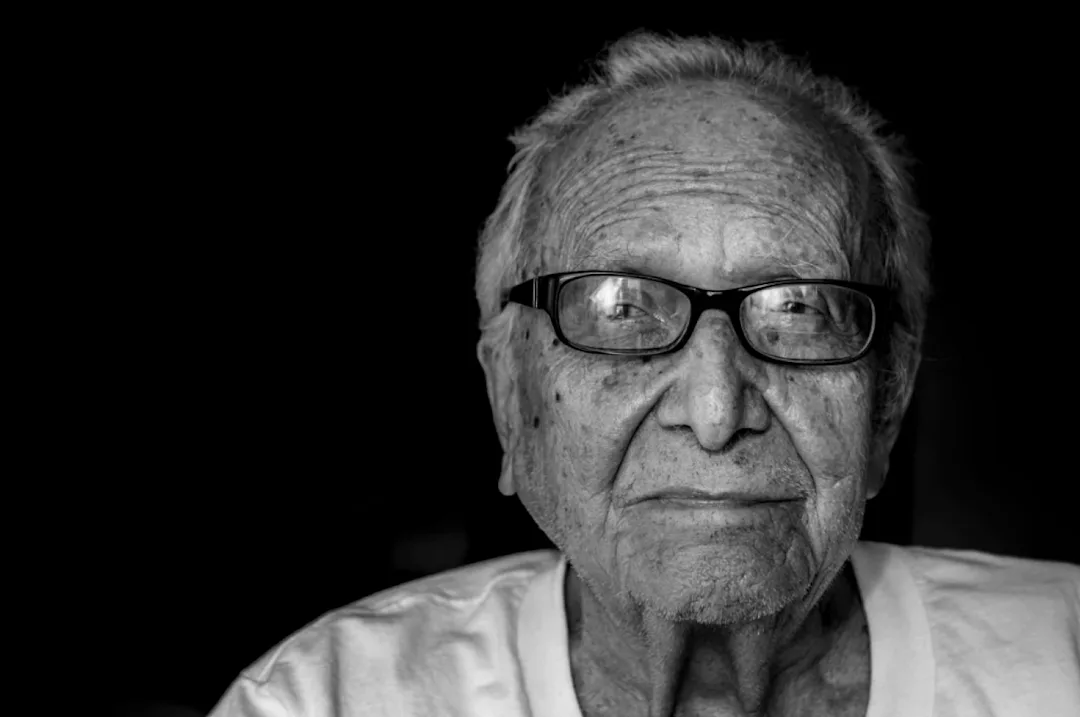 Close up black and white image of an old man looking straight on to the camera. His face is calm and confident