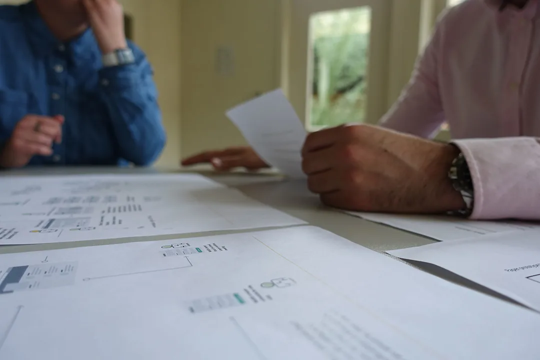 Zoomed in shot of the hands of two people sitting at a table. There are some printed diagrams on the table. 