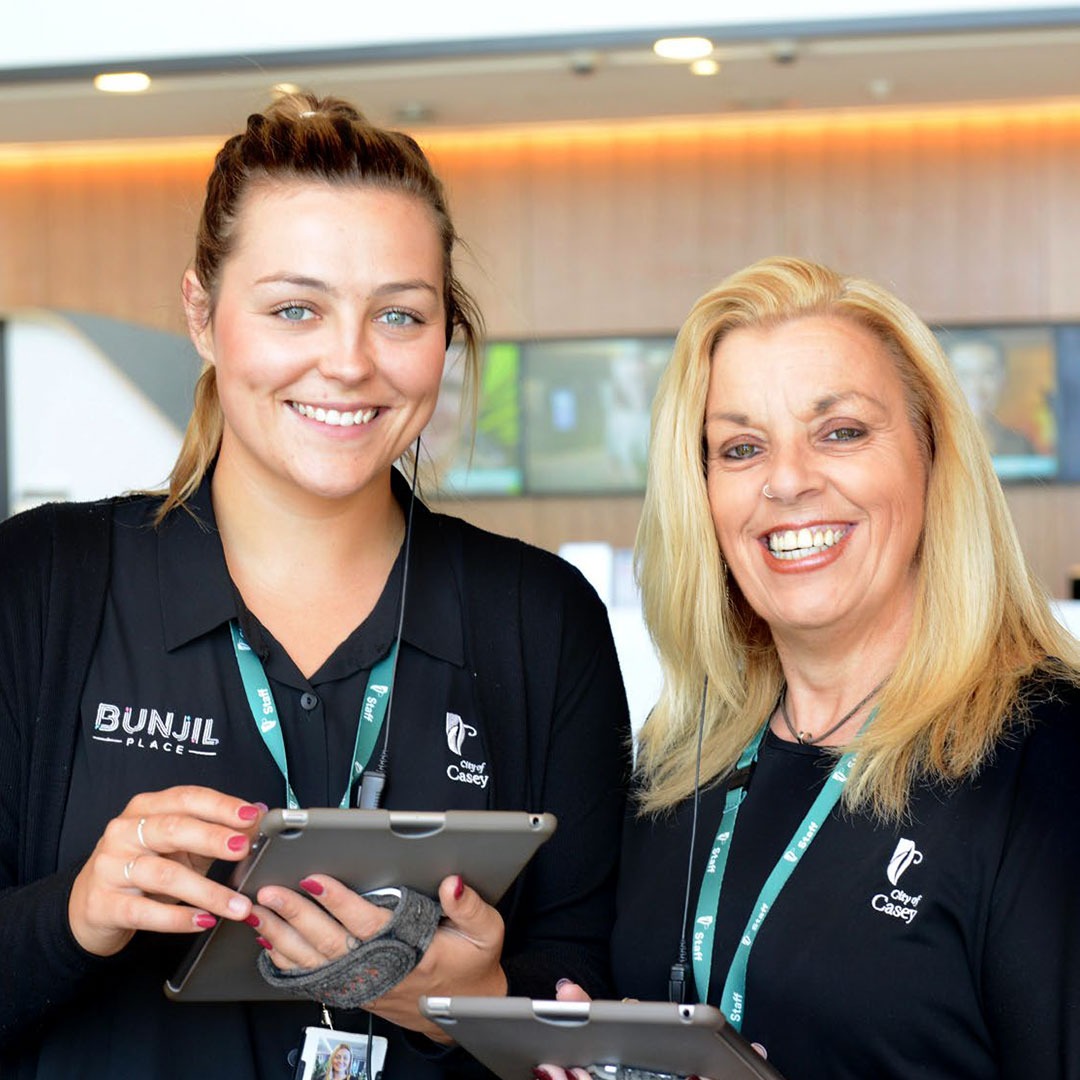 Two members of City of Casey staff supporting visitors to Bunjil Place with tablet devices.