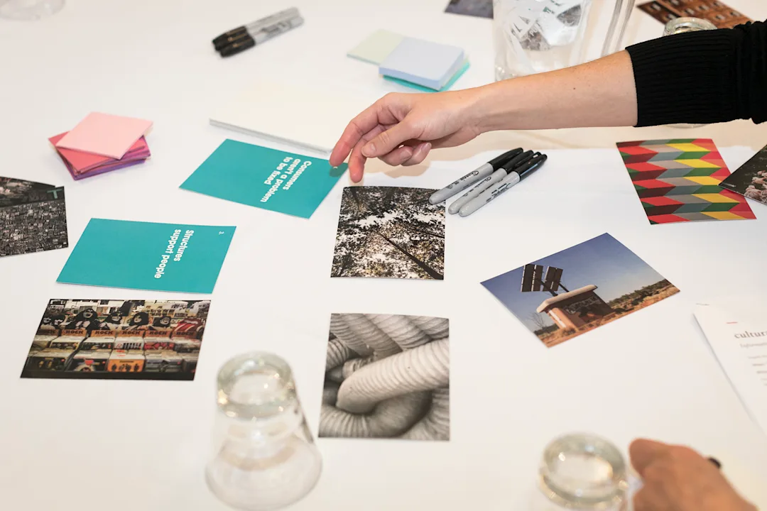 A photo taken during the co-design workshop showing a table with photos, post-it notes, pens and people's hands gesturing whilst they discuss ideas and thoughts.