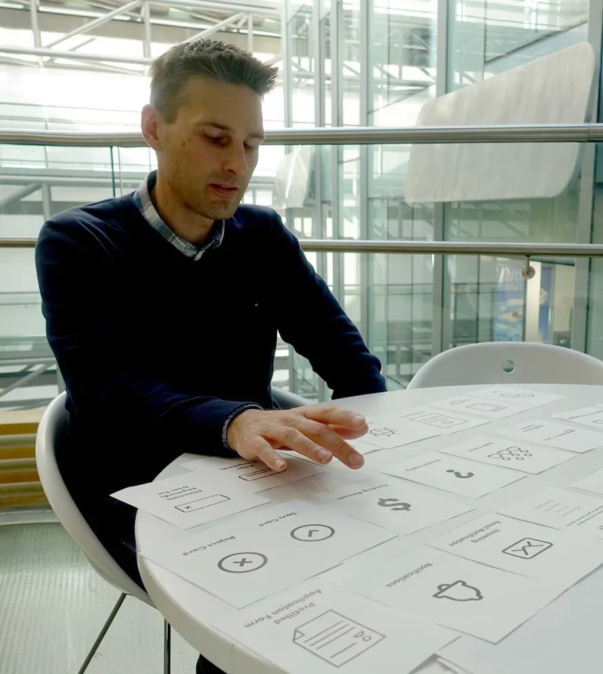 A man sitting at a table doing a card sort. Cards cover the table, and each card has a product feature or detail, such as notifications, funding amount and pre-filled forms. He is looking at the cards and choosing which ones are most valuable to him.