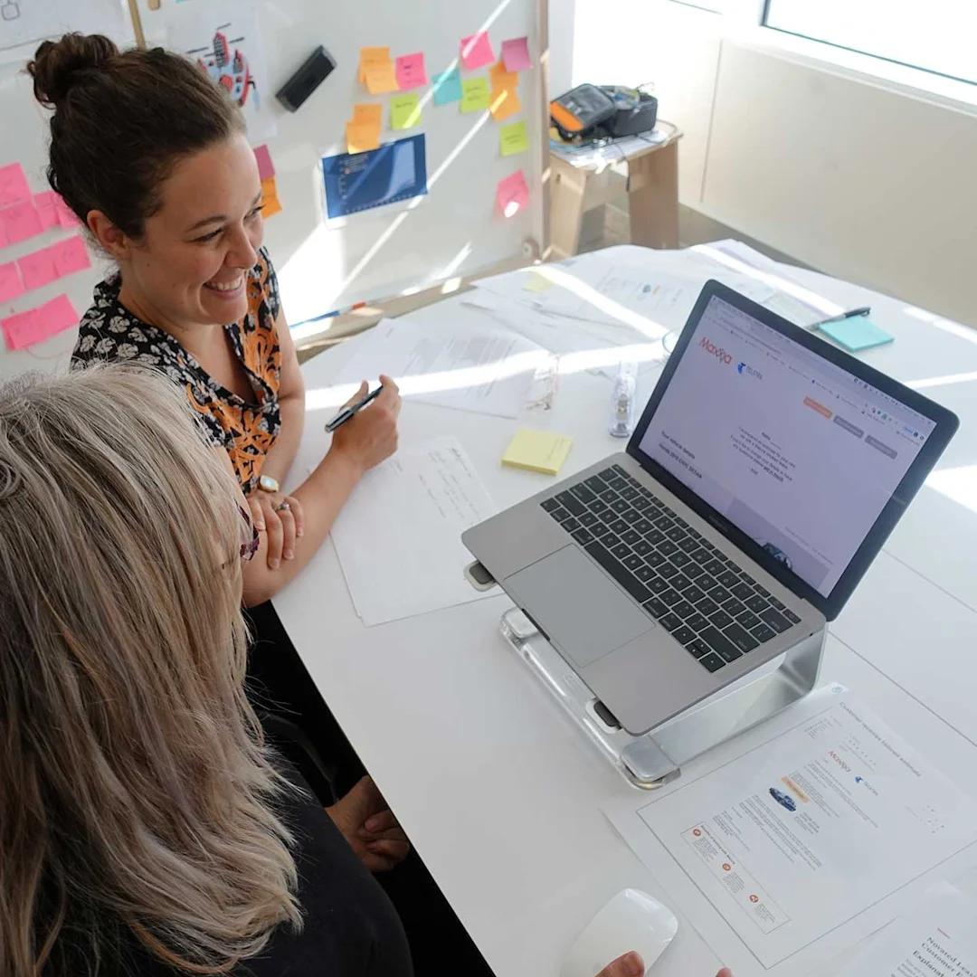 Two people are sitting at a table looking at a laptop. A website is shown on the laptop screen. One of the people is taking notes while the other person looks at the website.