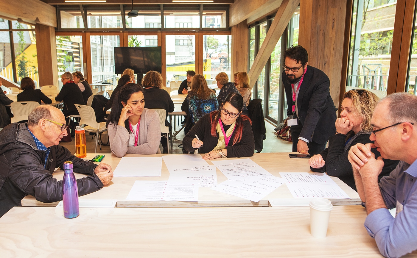 A table of five participants at a workshop being facilitated by Reuben. They are looking towards one participant who is taking notes. 