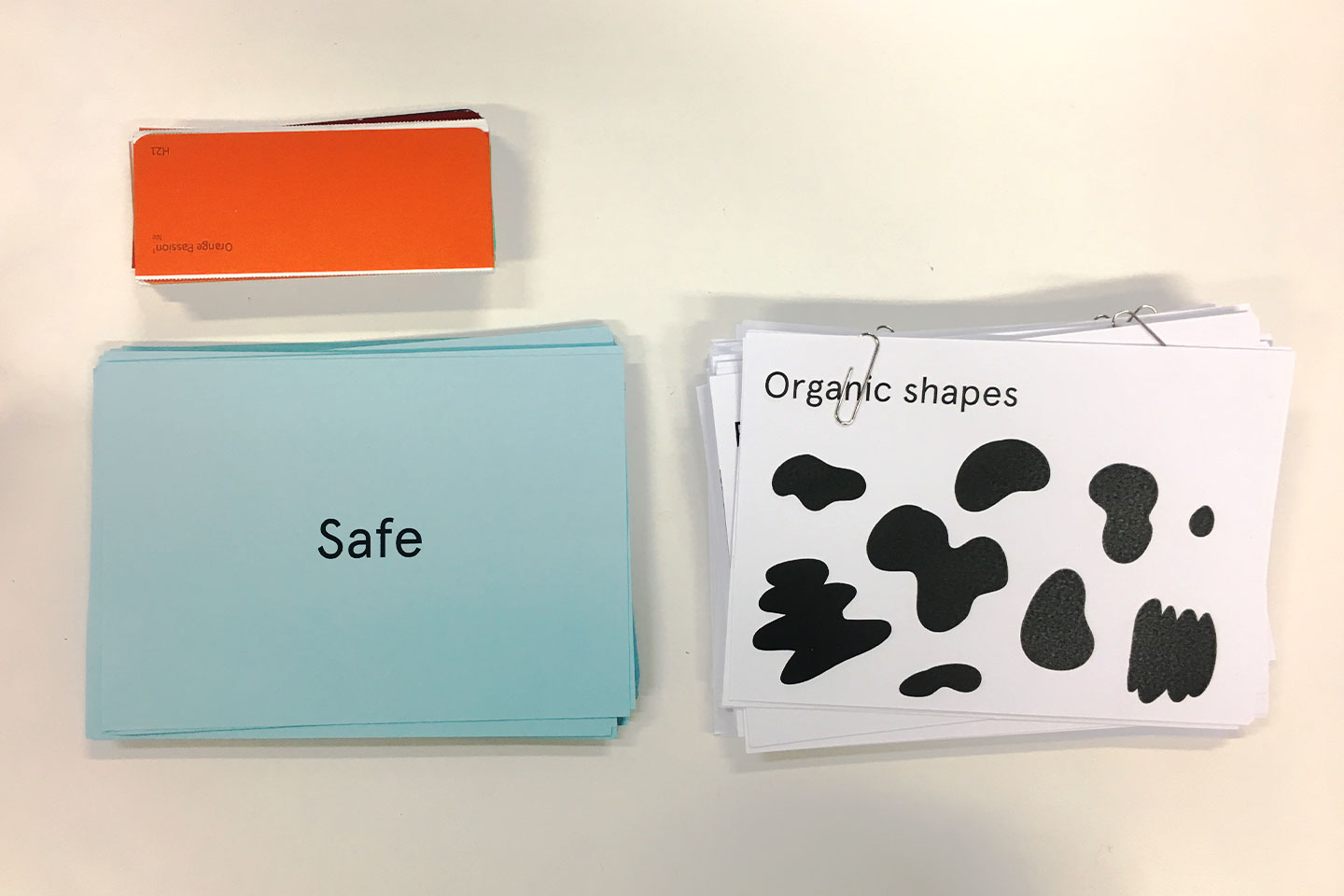 A photo of some sample branding workshop assets. There are three piles of cards. The first pile includes colours, the top card is orange. The second piles includes keywords, the top card says 'safe'. The third pile includes graphic elements, the top card says 'organic shapes' and have rounded shapes printed on it.  