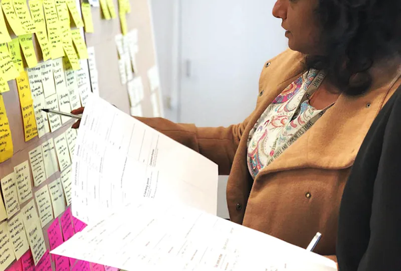 A photo of two people, looking at a wall of post-its notes, with worksheets in their hands.