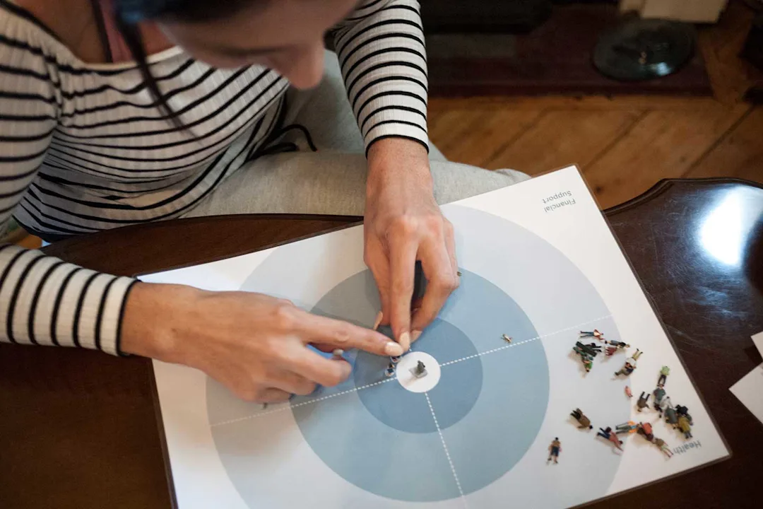 A woman sits at a table with a handful of small figurines and a sheet of paper. The woman uses the props to help tell her story of the death of a loved one. 