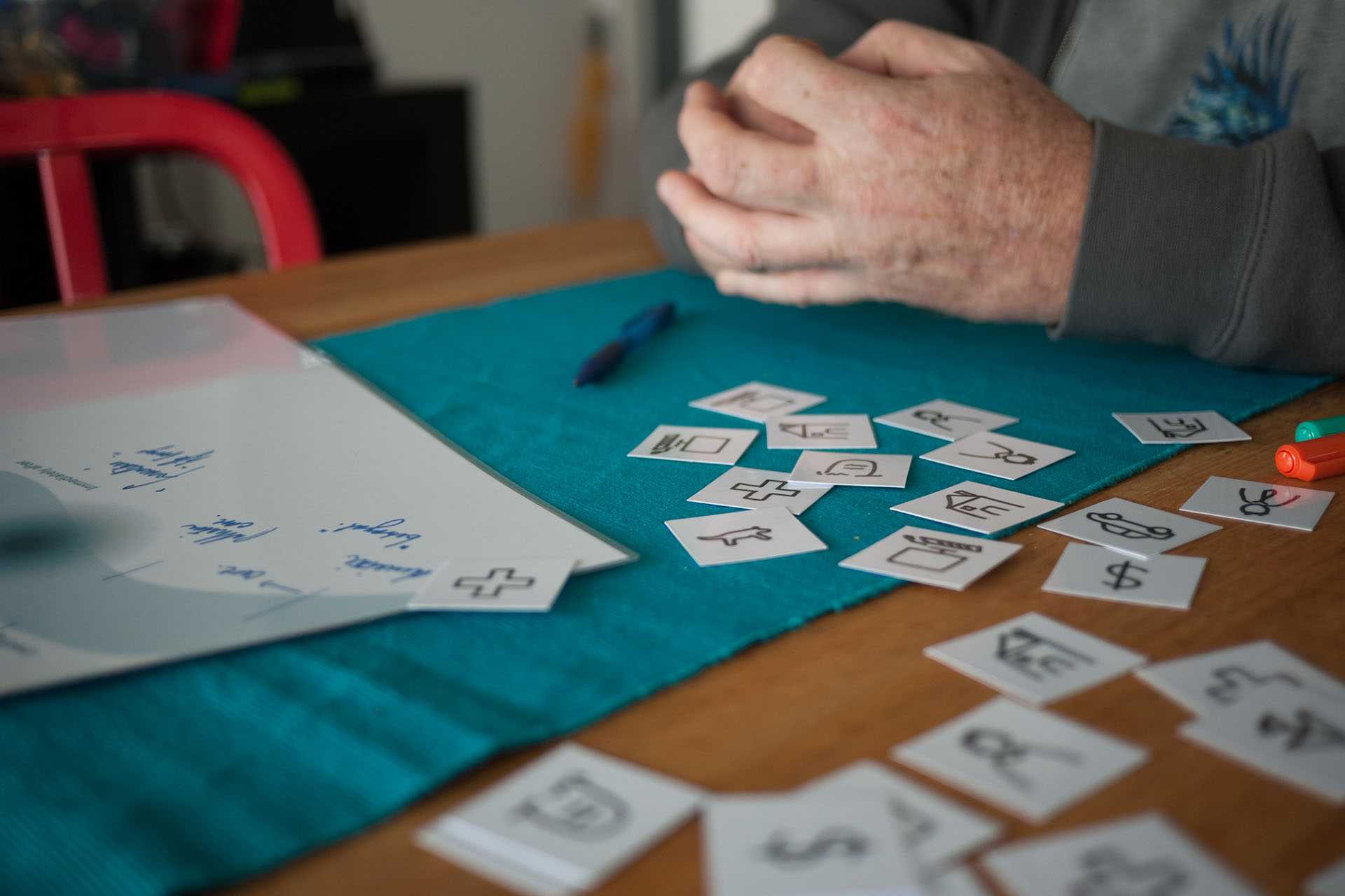 A photo zoomed into part of a dining room table, where a person sits alone. We can see their hands only. Small cards with printed icons are scattered across the table. These cards are printed with symbols for money, pets, people, health and more. 