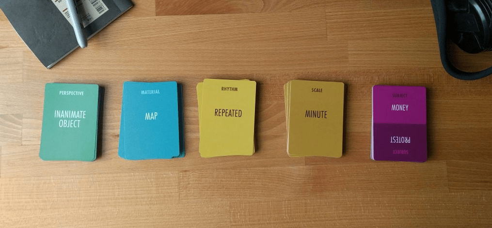 5 stacks of cards from the 'More Than Research' Game laid out on a table next to a notebook and pen. The top cards read "Perspective: Inanimate Object", "Material: Map", "Rhythm: Repeated", "Scale: Minute", and "Subject: Money".