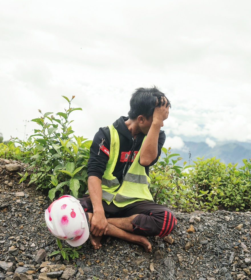 A man sits on the edge of a mountain slope, looking out over the clouds below. He is wearing a high-vis jacket and has a hard hat next to him. 