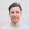 A photo of Jack Fahy smiling. He's outside in front of brick wall and it's a bright day.