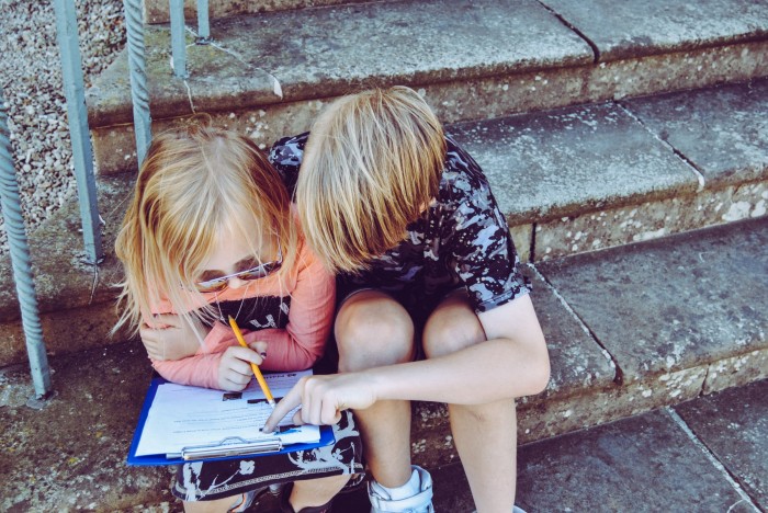 Two blonde children look at a clipboard while sitting on stairs outside