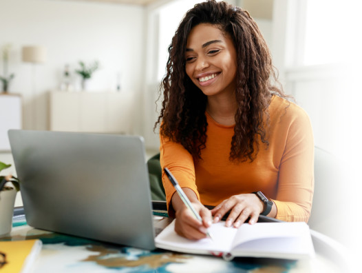 Woman sitting at a desk working on a laptop and taking notes in a notebook